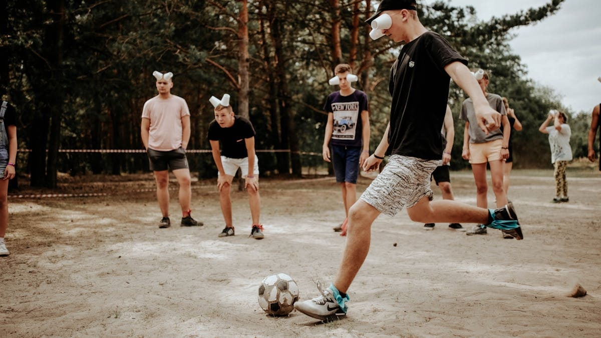 Group of teenagers wearing masks playing a soccer game in a sandy outdoor setting.